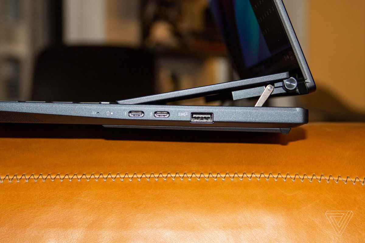 Asus Zenbook Pro 14 Duo, kahverengi bir kanepede sol tarafa doğru açılı.  Her iki ekran da Asus Zenbook Pro OLED logolu çok renkli bir arka plan gösteriyor.