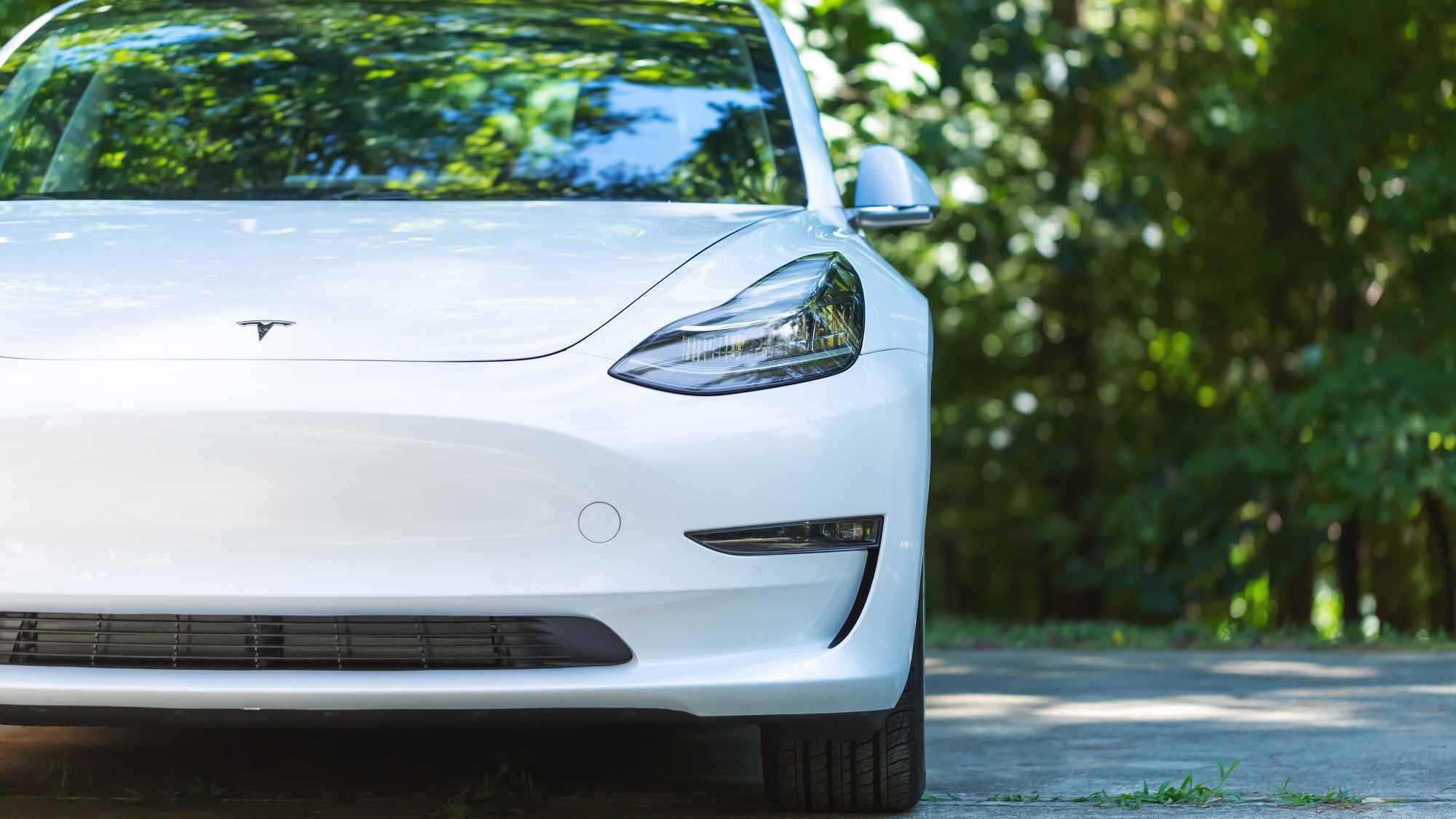 Güneşli bir günde arka planda ağaçları olan çimentolu yolda beyaz renkte tamamen elektrikli bir Tesla Model 3.
