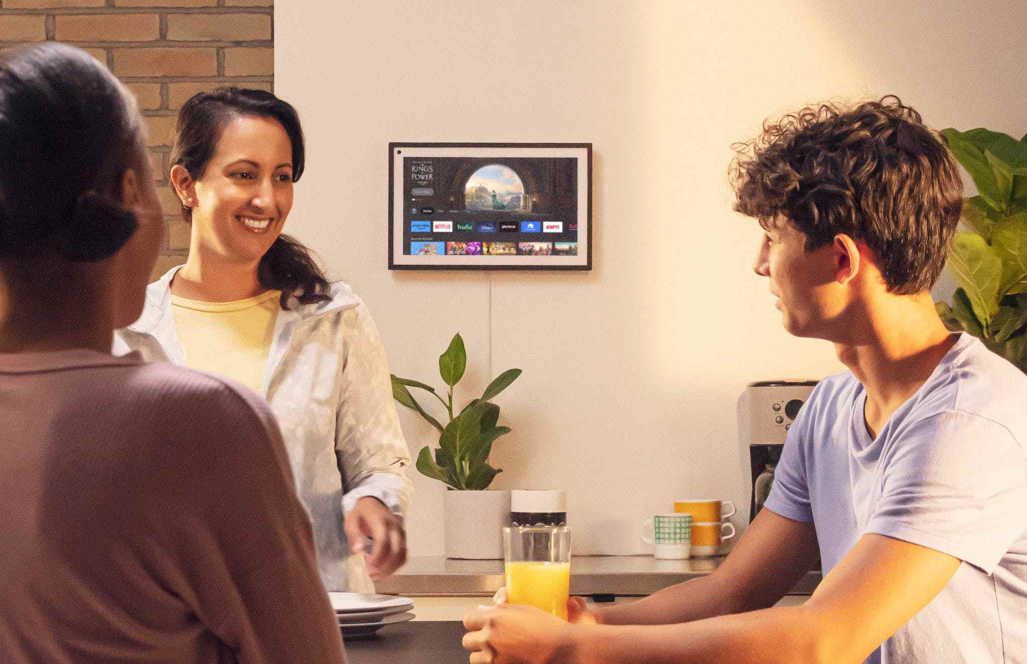 Amazon'un Fire TV işletim sistemini çalıştıran bir duvara monte edilmiş bir Echo Show 15'in görüntüsü.  İnsanlar ön planda sohbet ediyor.