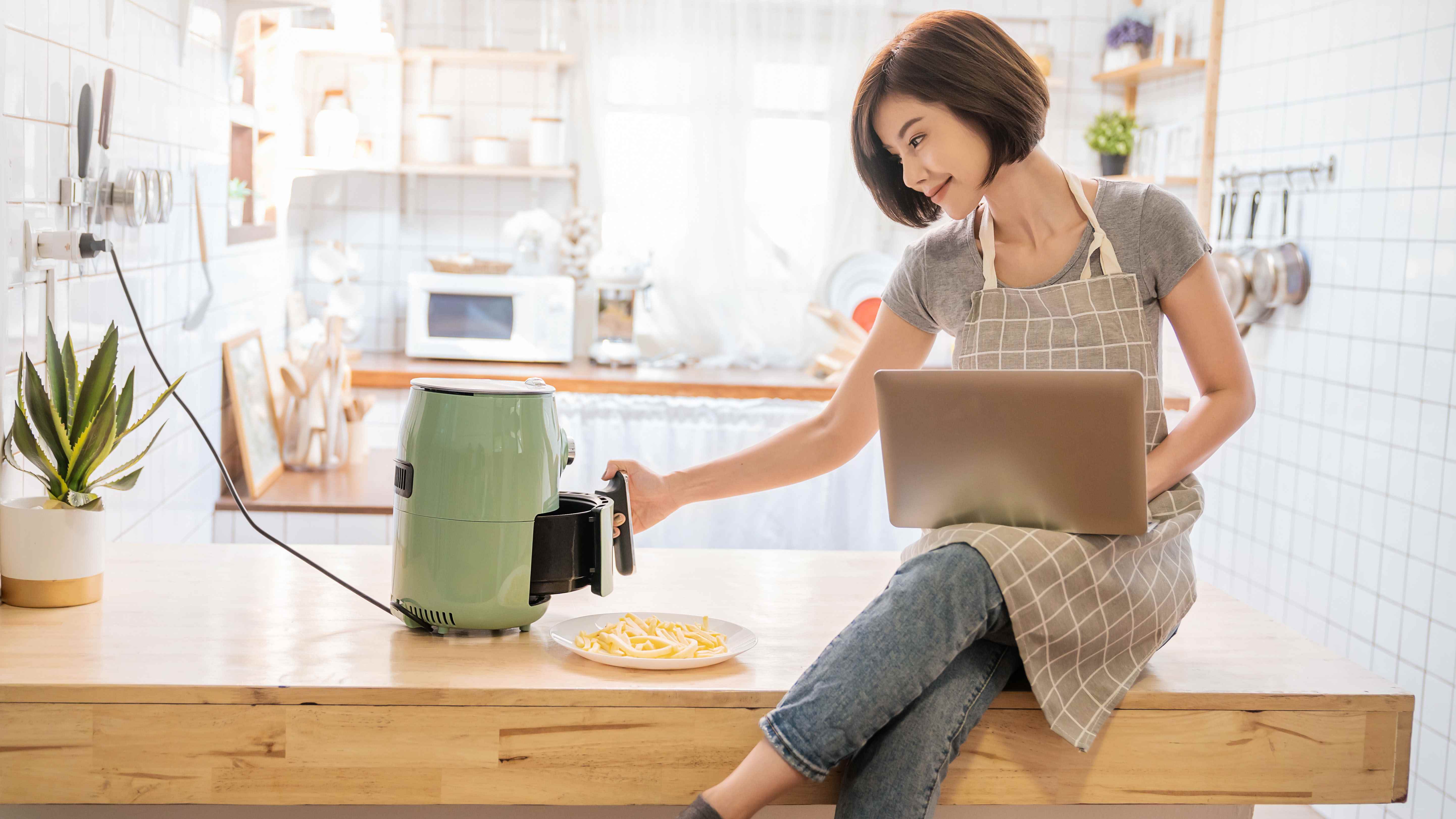 Bir tezgahta otururken kucağında dengeli bir dizüstü bilgisayar ve hava fritözüyle yemek pişiren bir kadın