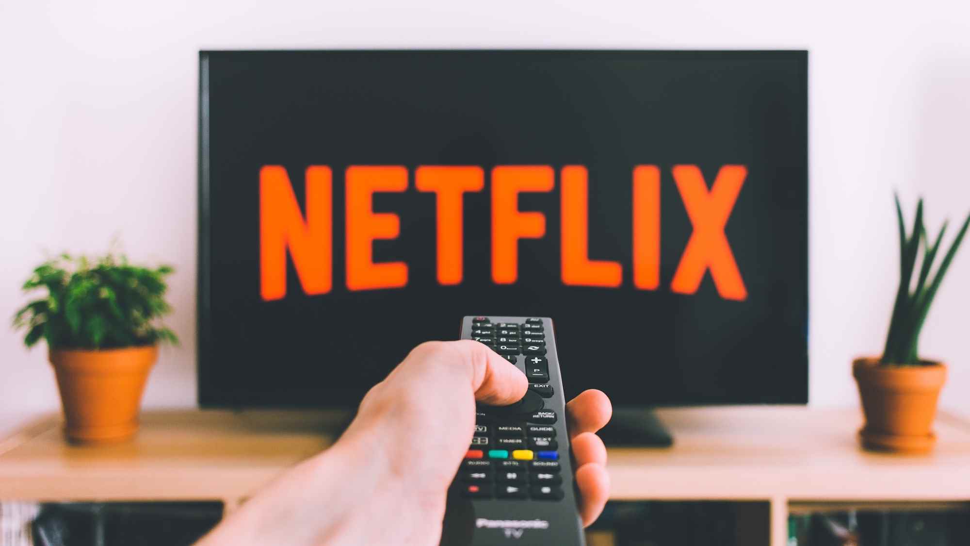 Netflix logolu bir TV, uzaktan kumandayı tutan bir elin arkasında oturuyor