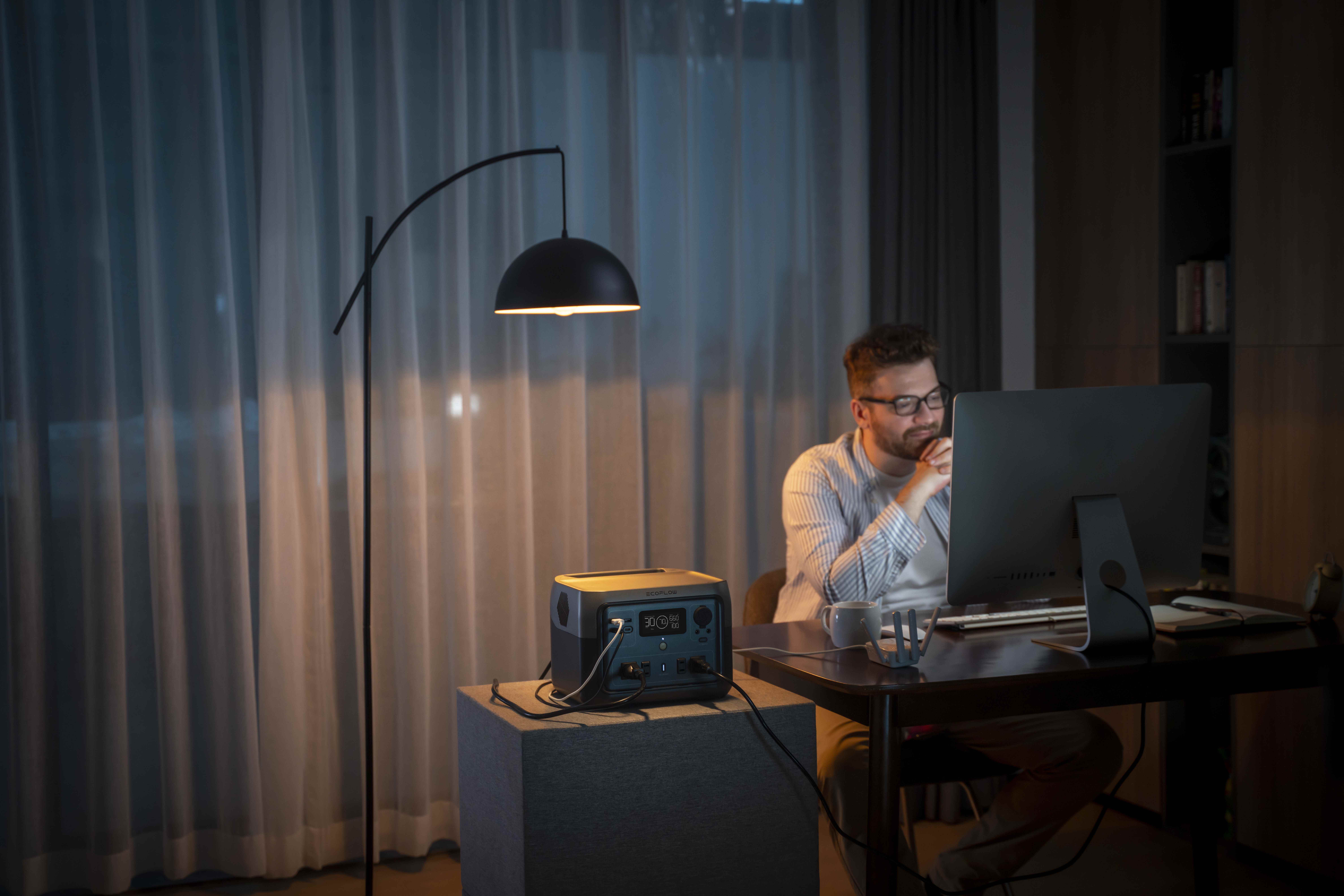 Bir adam ofisinde bir lambanın ışığı altında otururken bir River 2 Max pili bir masaüstü bilgisayara ve aksesuarlara güç sağlar.