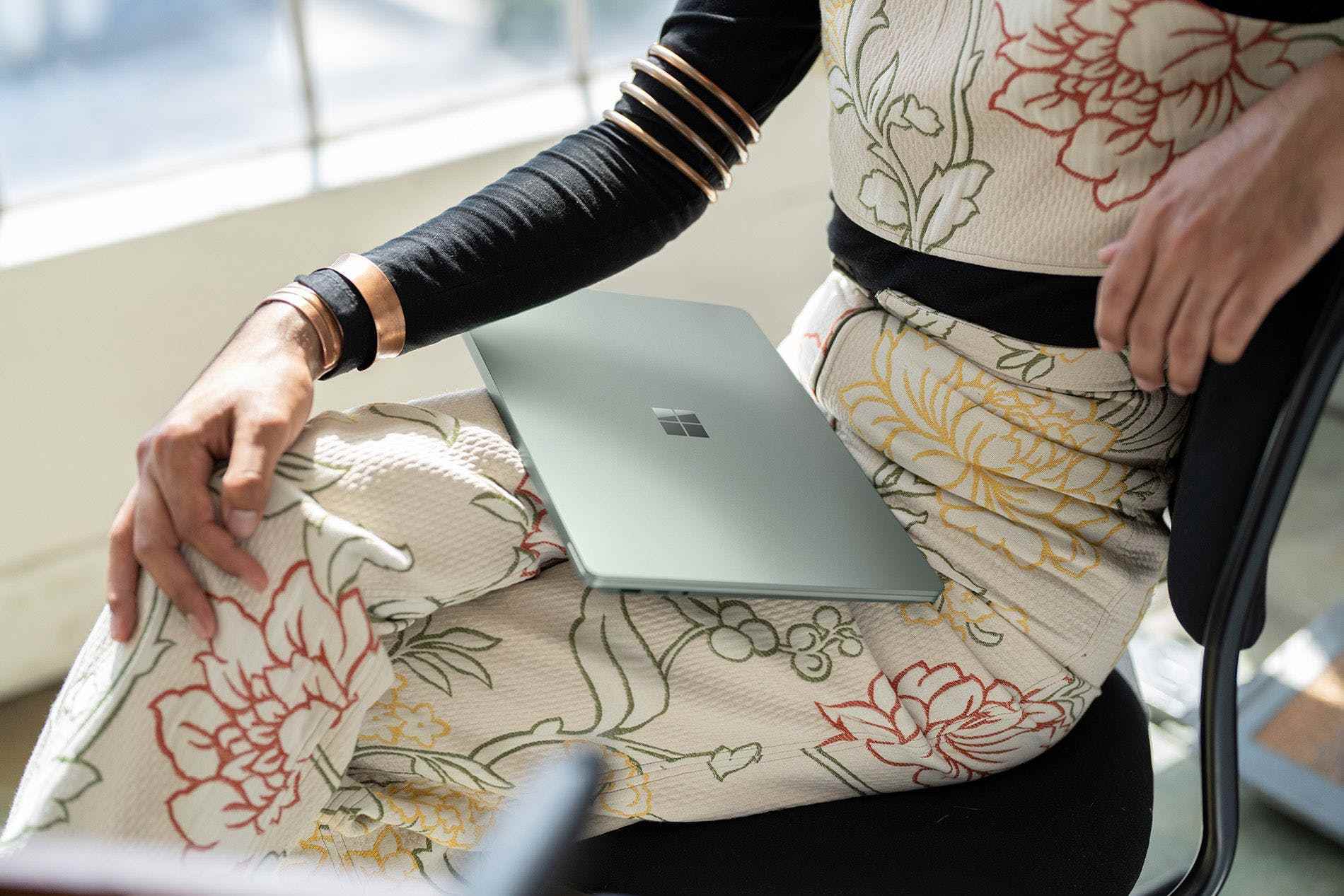 Çiçekli bir elbise içinde oturan bir kullanıcı, Microsoft logosunun göründüğü kapalı bir Surface Laptop 5'i kucağında tutuyor.
