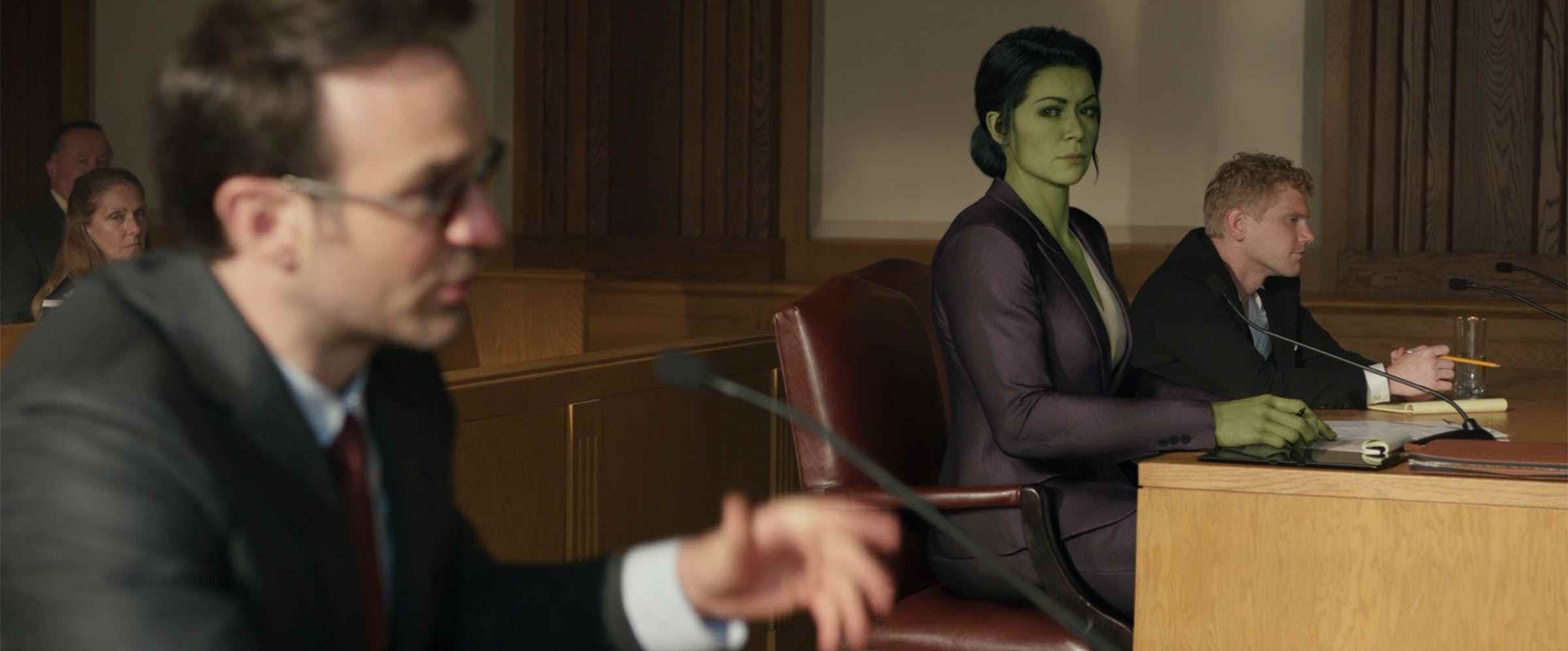 Mahkeme salonunun önünde müvekkilinin yanında oturan mor takım elbiseli yeşil bir kadın, takım elbiseli sarışın bir adam eğildi.  Kadın başka bir avukata bakıyor, kırmızı gözlük takan bir adam.