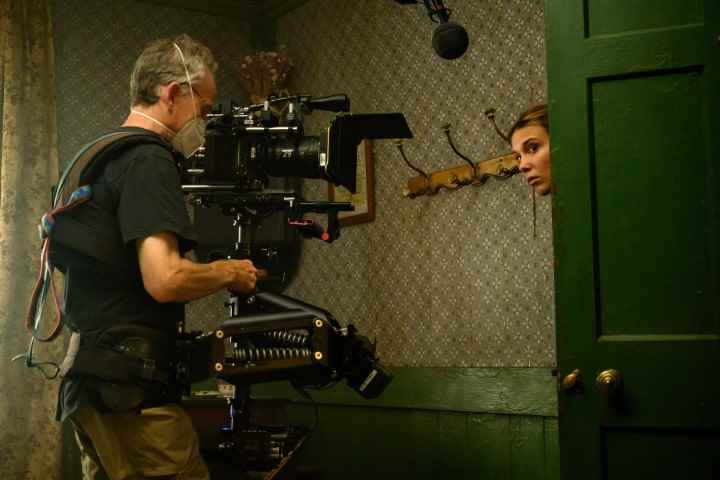 Millie Bobby Brown, Enola Holmes 2'nin kamera arkası fotoğrafında filme alınırken bir kapıdan içeri bakıyor.