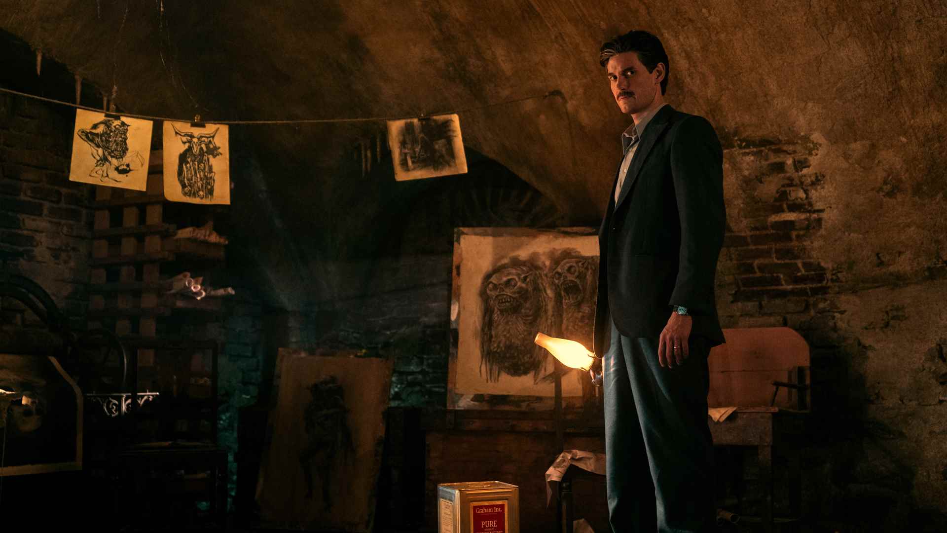 Will Thurber, ateşle aydınlatılmış bir odada, elinde bir fenerle, Netflix'teki Pickman's Model'deki bazı sanat eserlerinin yanında duruyor