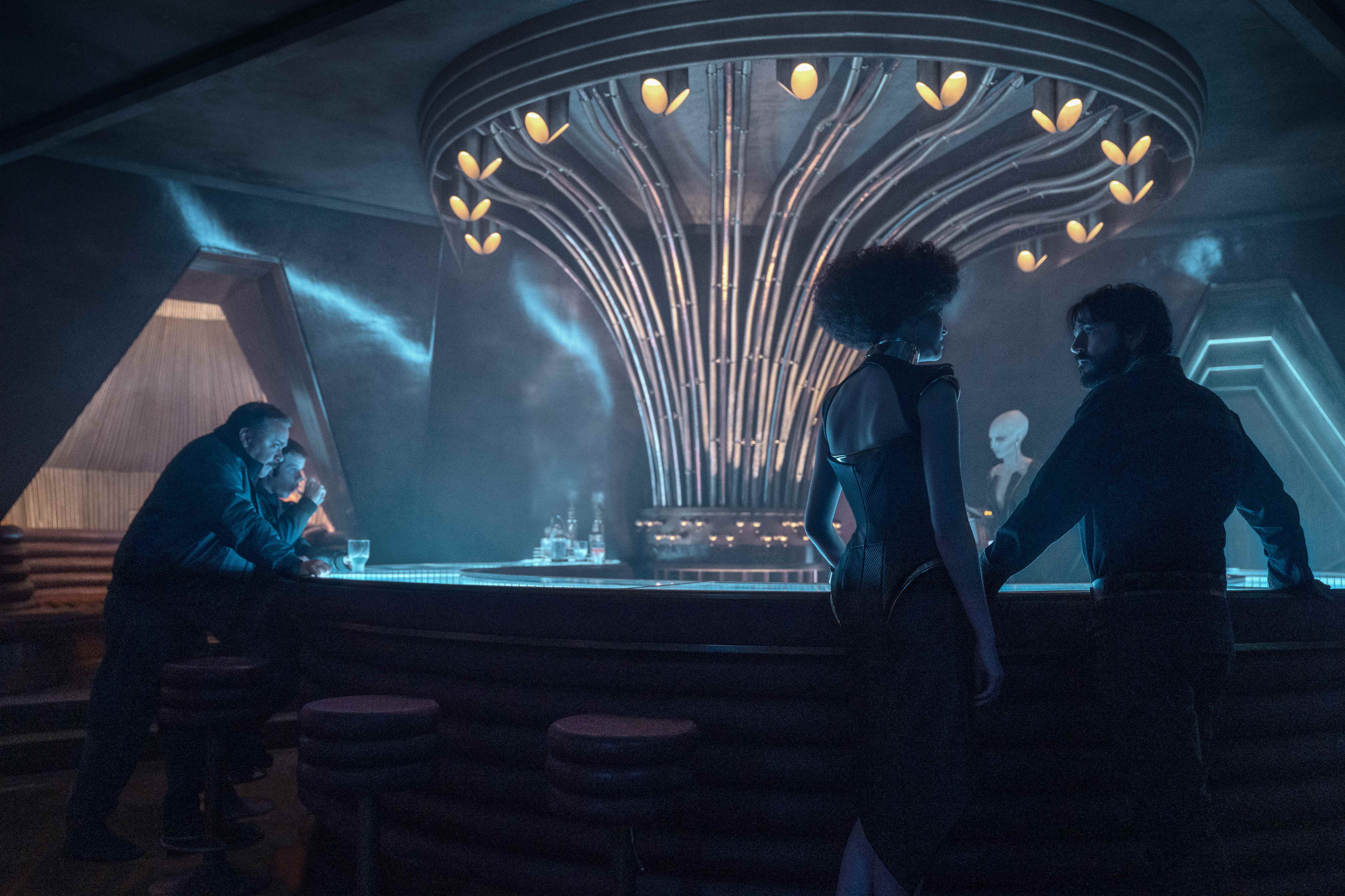 Andor'da Kravas (Lee Boardman) ve Verlo (Stephen Wright) bakarken Cassian Andor bir hostesle (Margaret Cline) konuşuyor.