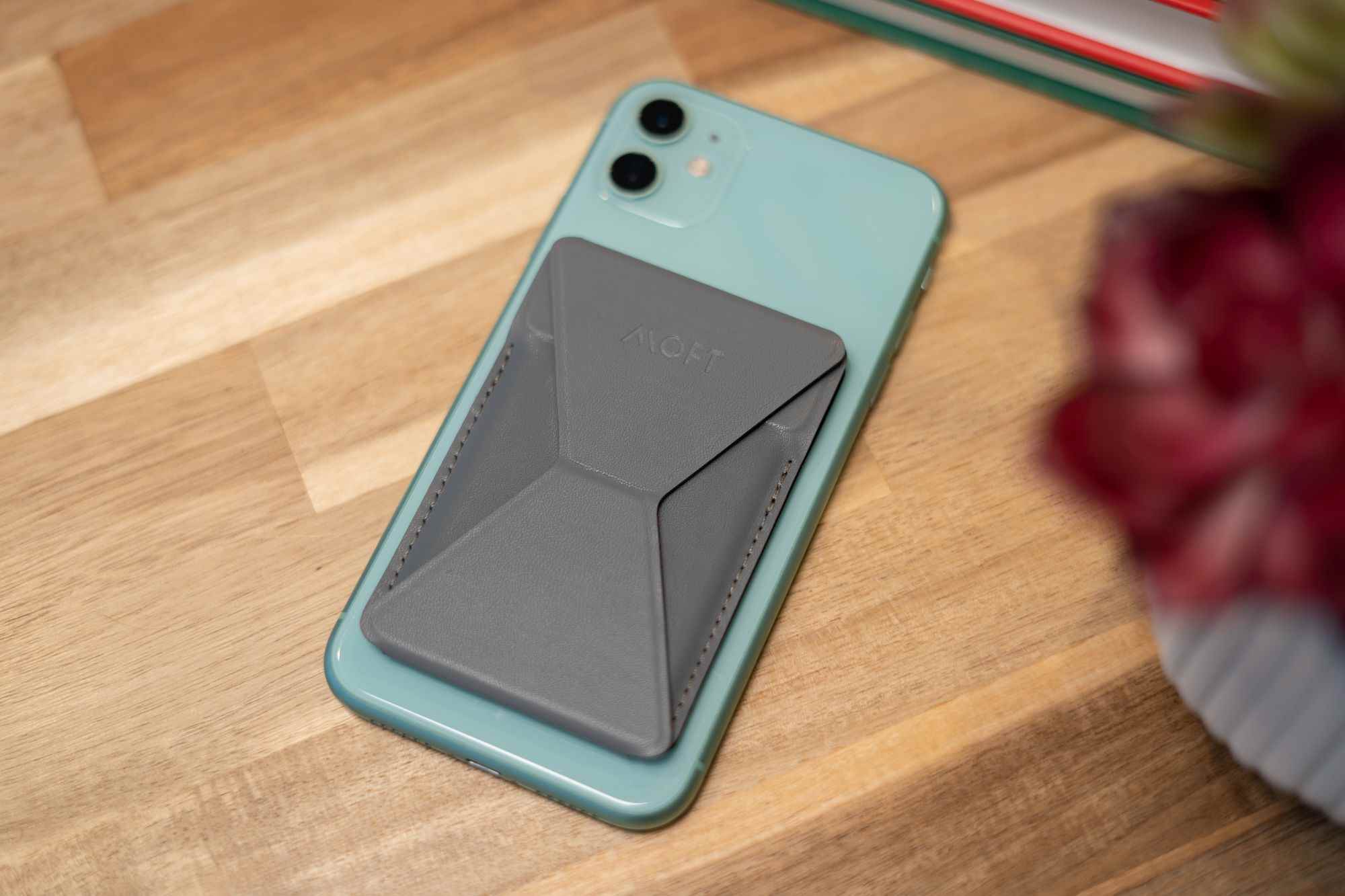 Yeşil bir iPhone 11'in arkasındaki Gri Moft cüzdan yüzü aşağı bakacak şekilde masaya.