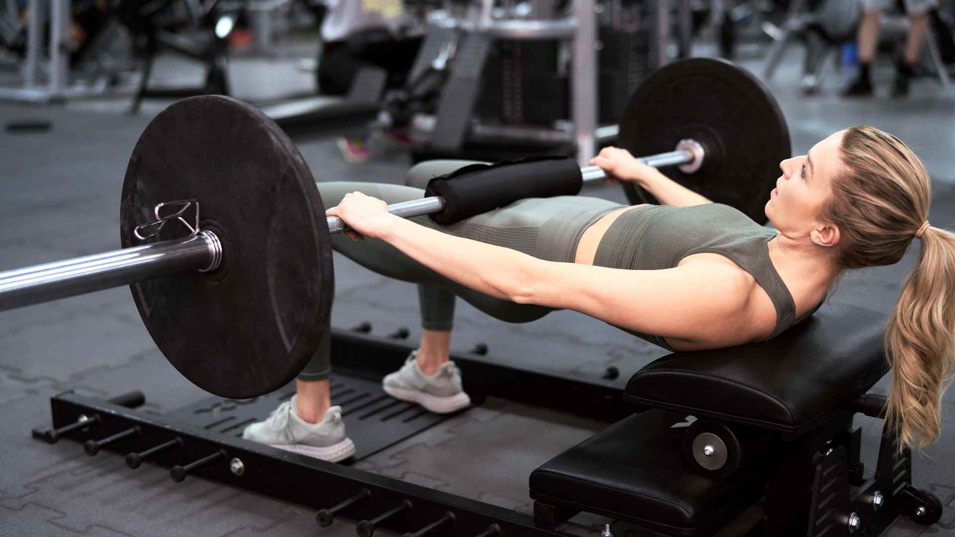 Spor salonunda bir bankta halterle kalça hareketi yapan kadın