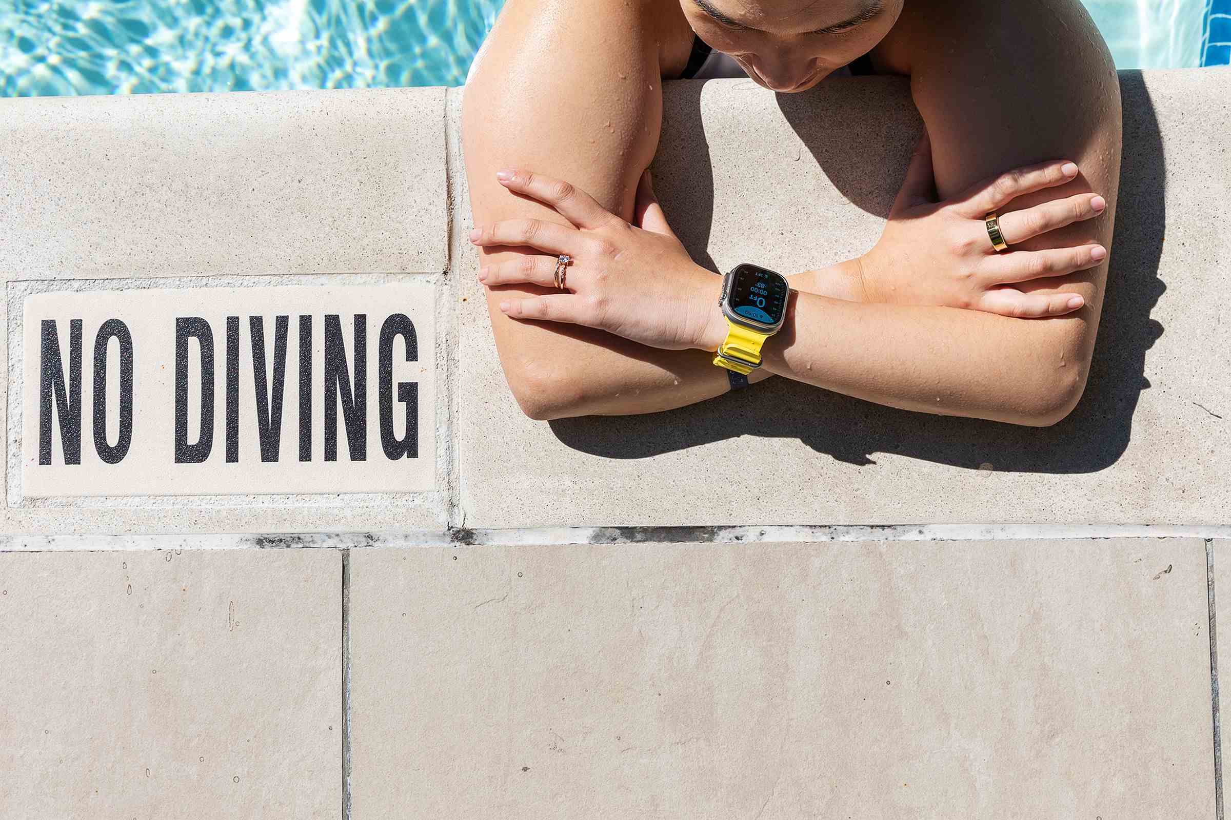 Havuzun kenarına eğilmiş kadın, dalış yasak levhasının yanında Apple Watch Ultra takıyor.