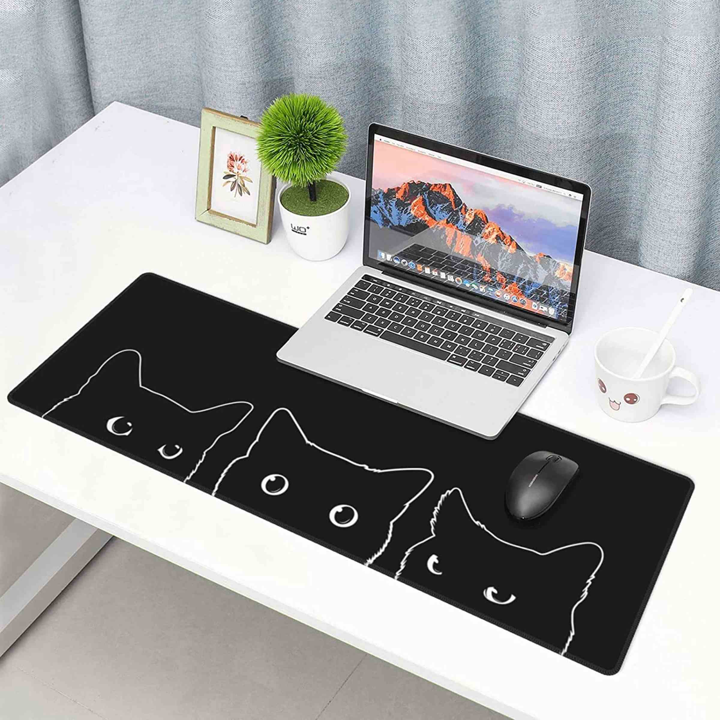Bir dizüstü bilgisayar, bir bitki ve küçük çerçeveli bir resim ile beyaz bir masa üzerinde üzerinde üç kedinin ana hatları olan uzun siyah bir fare altlığı.