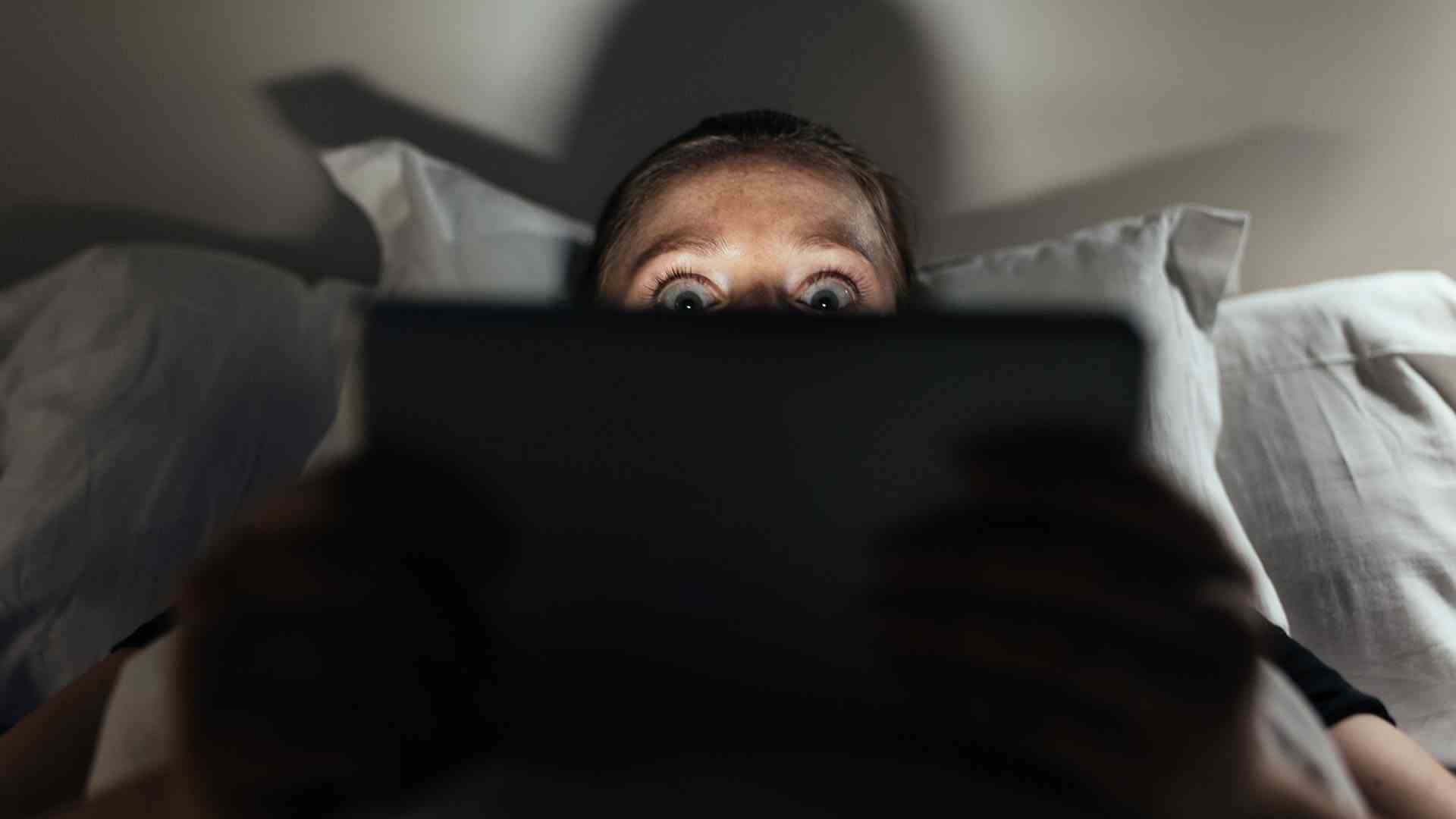 Kadın korkmuş görünüyor, yatakta dizüstü bilgisayar ekranında bir şeyler izliyor.