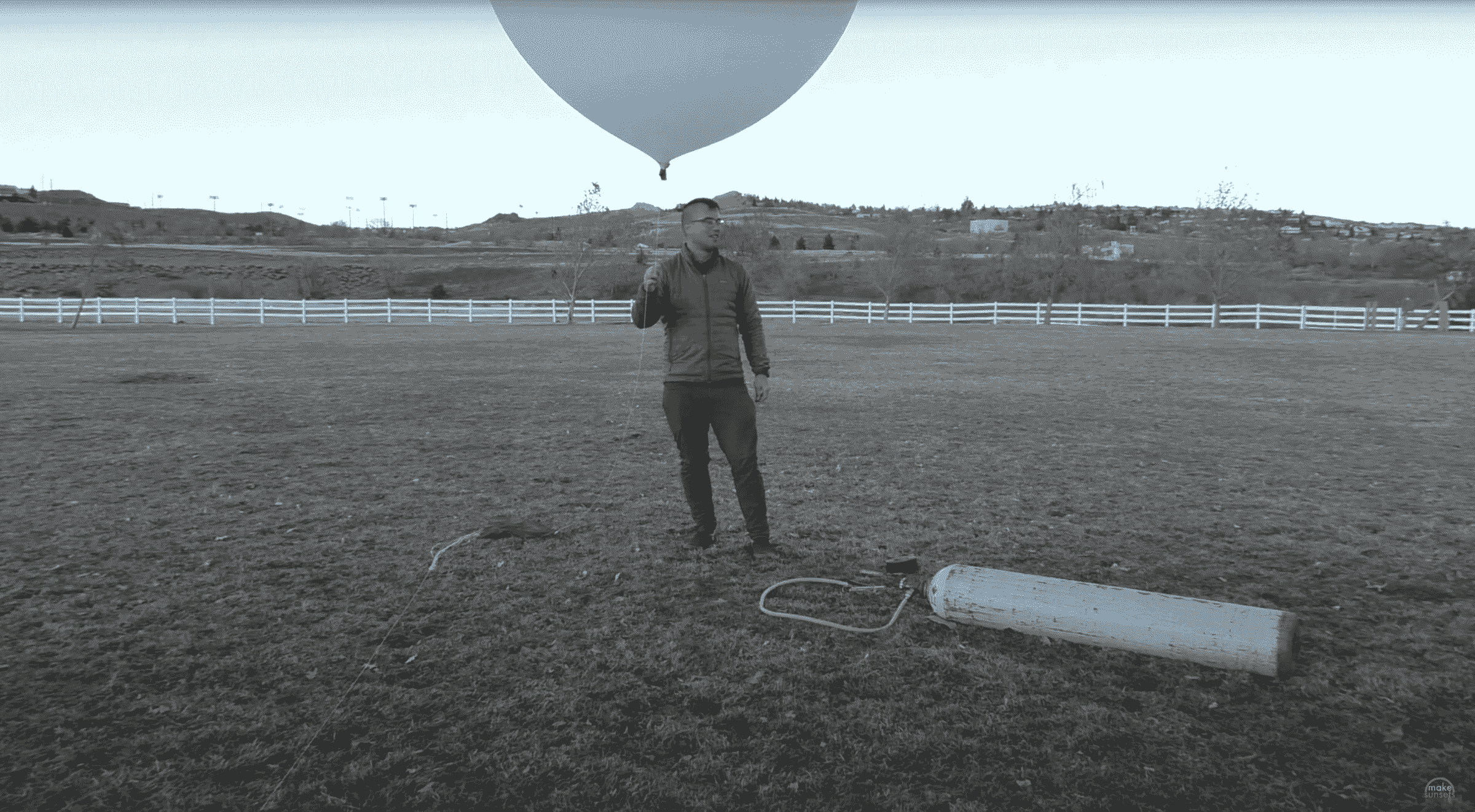 Bir adam büyük beyaz bir hava balonu taşıyan çimenli bir alanın ortasında duruyor.