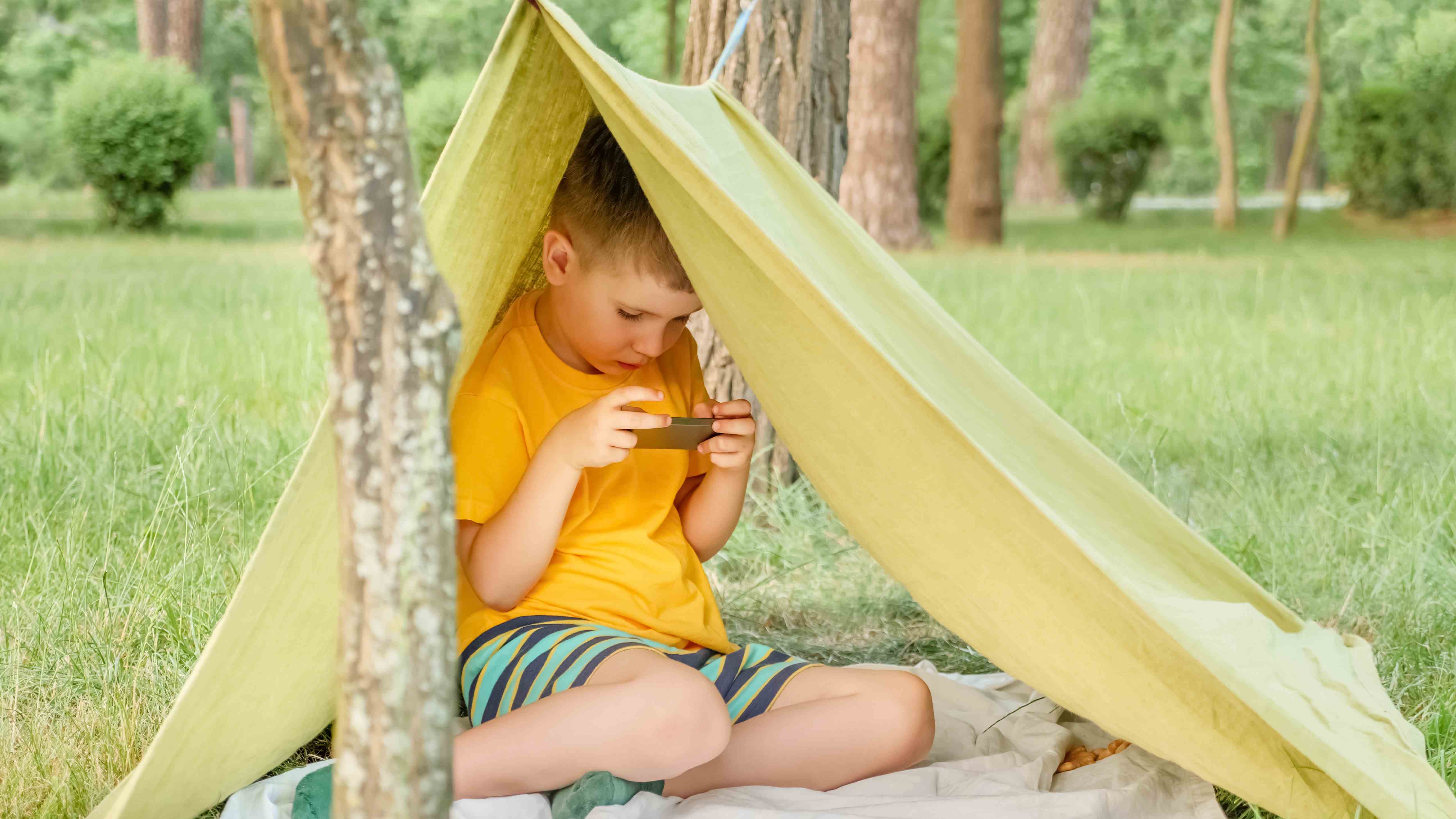 Ormanda derme çatma bir çadırda otururken telefonunda Netflix izleyen küçük bir çocuk