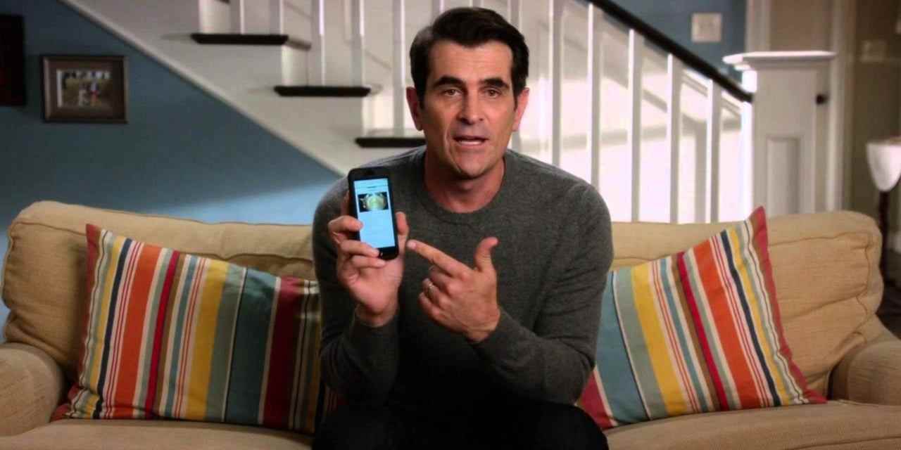 Phil kanepede oturmuş Modern Family'deki telefonunu işaret ediyor