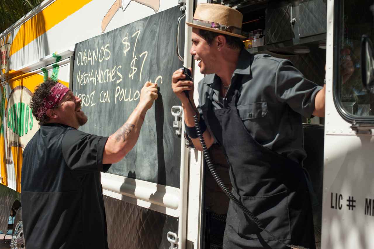 (Soldan sağa) Jon Favreau, Carl Casper rolünde bir yemek kamyonunun yan tarafında bir menü yazarken John Leguizamo, Martin Chef'de mikrofona konuşurken