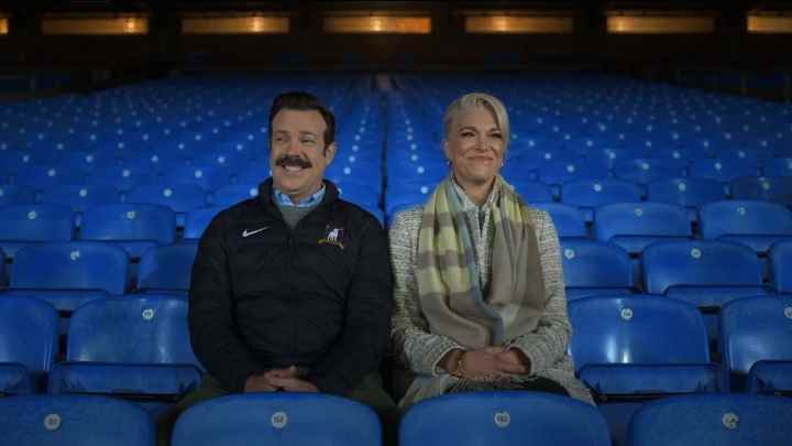 Ted Lasso'dan bir sahnede stadyum koltuklarında oturan Ted ve Rebecca birlikte gülüyorlar.